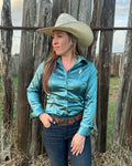 A woman wearing a shiny teal satin button-down shirt, dark blue jeans, a brown belt, and a beige cowboy hat stands in front of a rustic wooden fence outdoors.