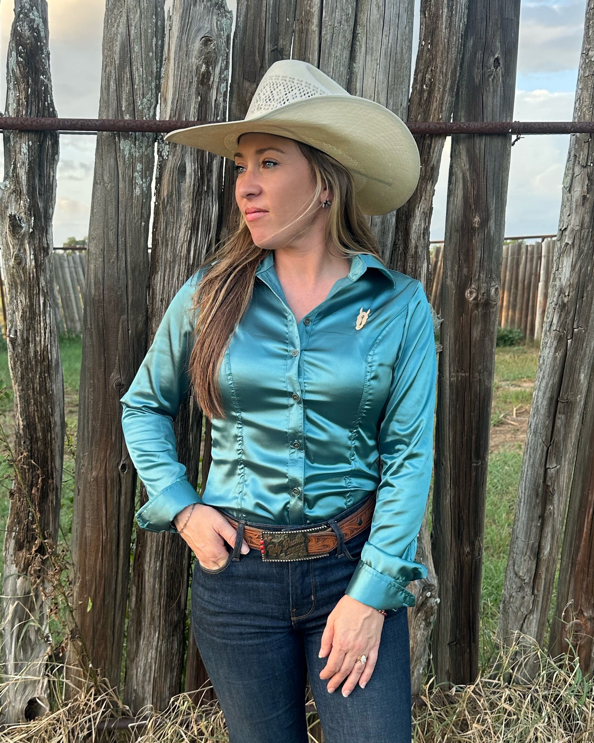A woman wearing a shiny teal satin button-down shirt, dark blue jeans, a brown belt, and a beige cowboy hat stands in front of a rustic wooden fence outdoors.