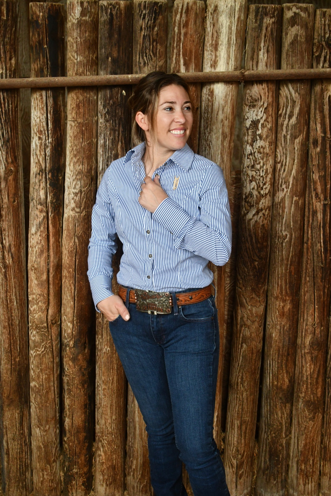 Woman wearing a blue pinstripe shirt and jeans standing against a wooden wall.