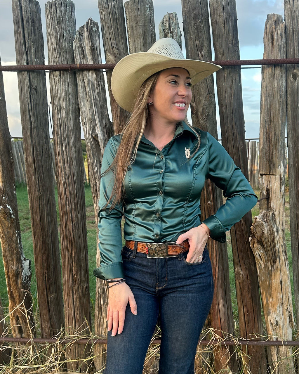 A woman wearing a dark green satin button-up shirt with a collared neckline, standing outdoors in front of a wooden fence.