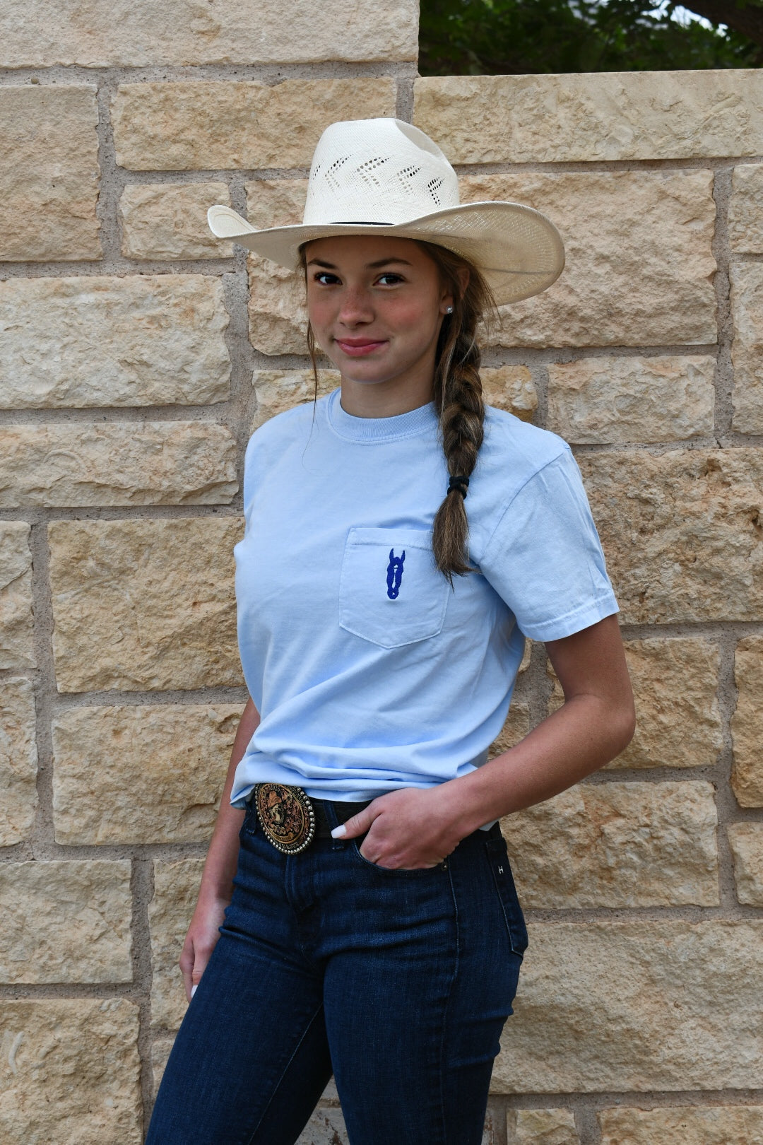 Person wearing a light blue shirt with a logo, dark pants, and a white cowboy hat against a stone wall.