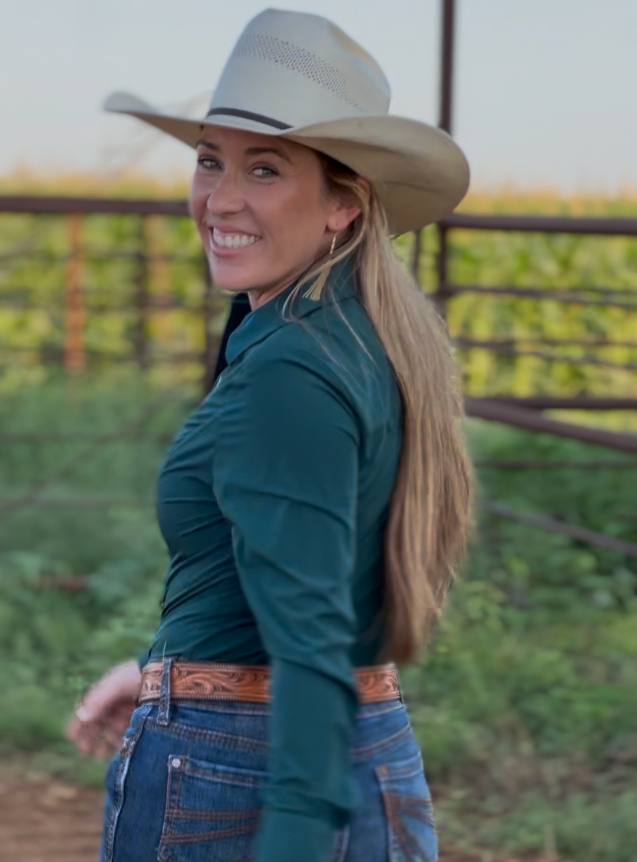 A woman wearing a light beige cowboy hat, a dark teal long-sleeve shirt, and blue jeans stands outdoors in front of a fence and field.