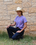 Person wearing a purple shirt and cowboy hat sitting against a stone wall.