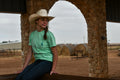 Person wearing a cowboy hat and green shirt sitting under a stone archway with hay bales in the background.