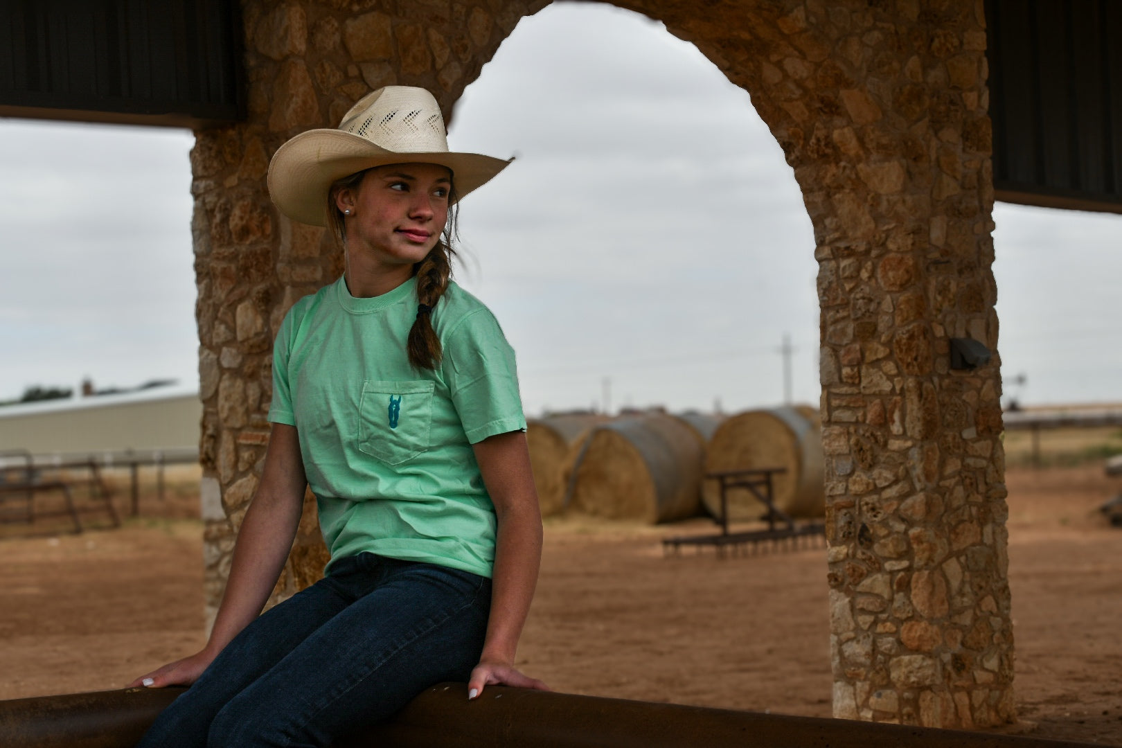 Person wearing a cowboy hat and green shirt sitting under a stone archway with hay bales in the background.